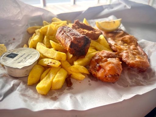 A fish and chips meal on an open paper wrapping featuring a small container of tartare sauce, a deep fried dim sim, a calamari ring, and a battered scallop. The chips and fried fish fillet have a wedge of lemon and also a sprinkling of chicken salt. 